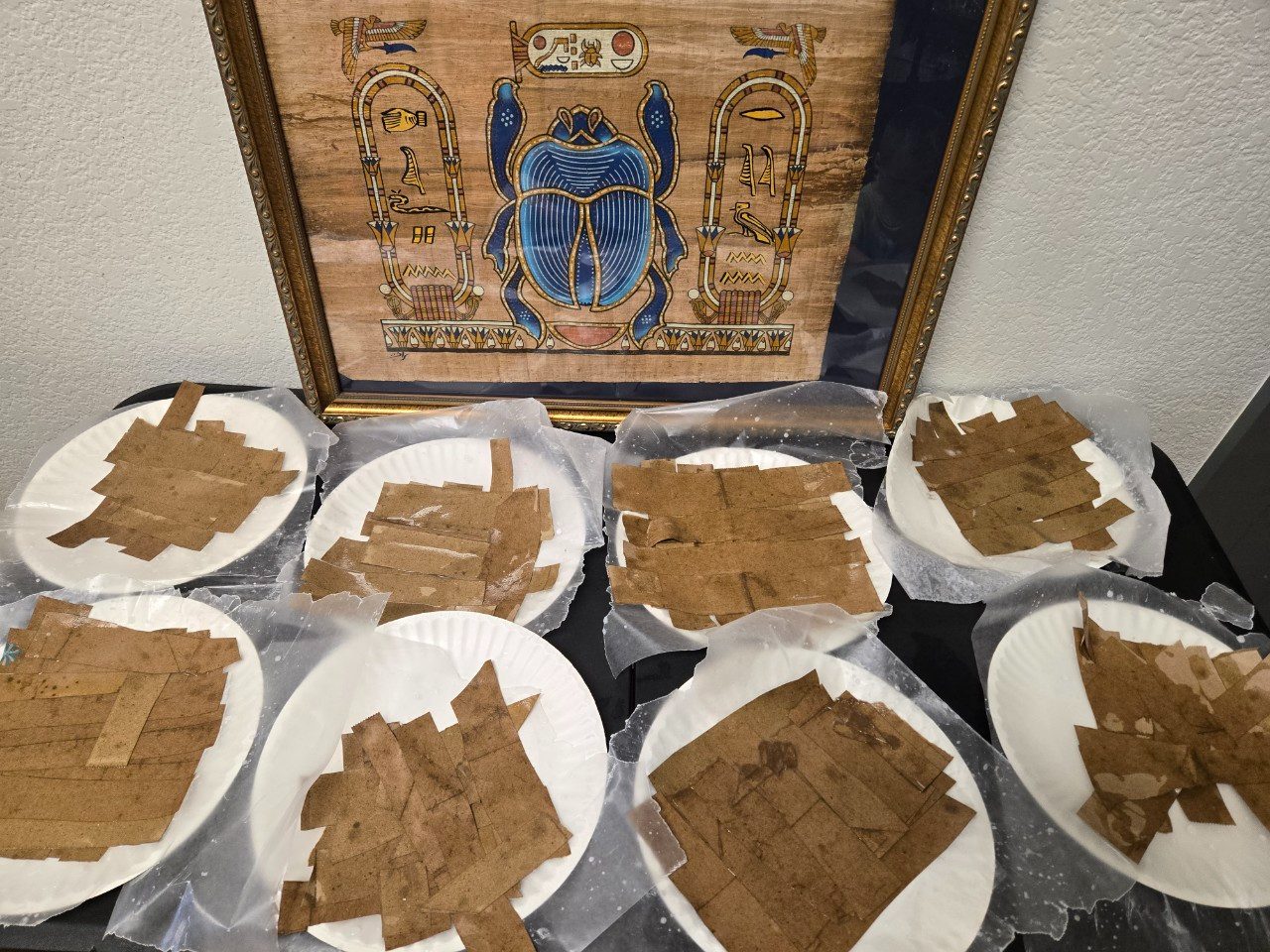 Handmade papyrus paper squares drying on plates beside an Egyptian scarab print